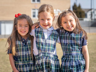 Three young girls in plaid school uniforms smile and hug outdoors, standing on grass with a building in the background. Bright, sunny day.