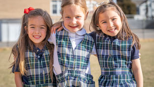 Three young girls in plaid school uniforms smile and hug outdoors, standing on grass with a building in the background. Bright, sunny day.