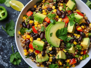 Quinoa salad in a bowl with avocado, black beans, corn, red peppers, cilantro; lime and jalapeño slices nearby on black marble.