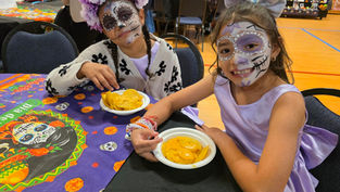 Two children in Day of the Dead face paint enjoy nachos at a festive event. One wears a floral crown. Colorful decorations are visible.
