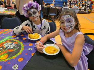 Two children in Day of the Dead face paint enjoy nachos at a festive event. One wears a floral crown. Colorful decorations are visible.