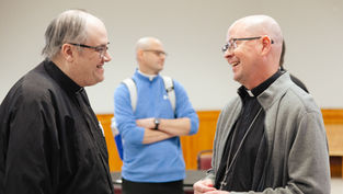 Two Catholic priests smile and talk together in a meeting room, with another man standing in the background.