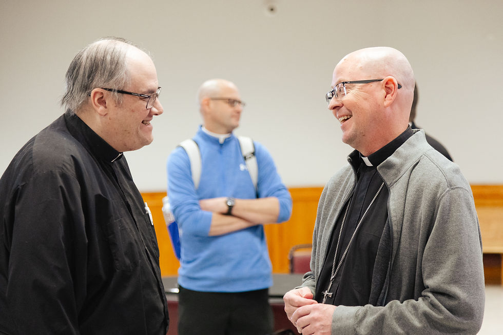 Two Catholic priests smile and talk together in a meeting room, with another man standing in the background.