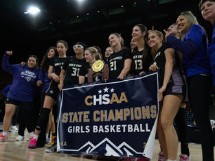 Girls' basketball team celebrates championship win, holding trophy and "State Champions" banner in stadium. Team wears black uniforms.