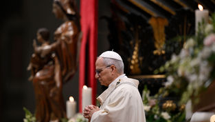 Cleric in white robe and cap prays with folded hands. Background shows a statue, red drape, and lit candles, creating a solemn ambiance.