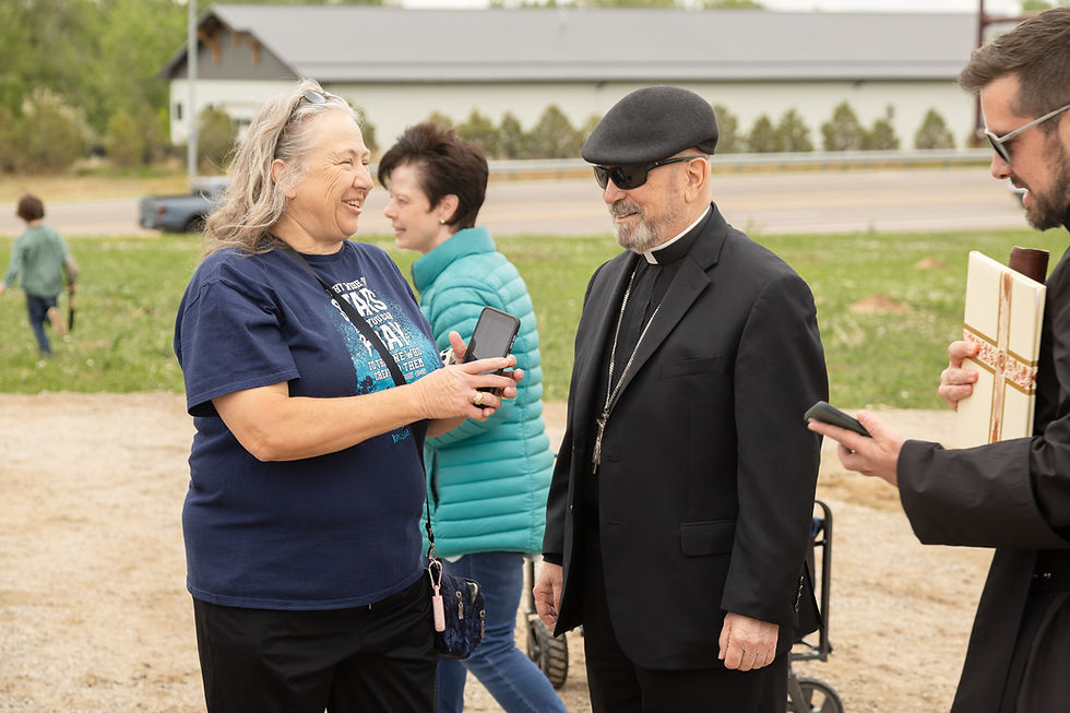Woman in navy shirt smiling and showing phone to a man in black suit and sunglasses outdoors. Others are near, one holding a document.
