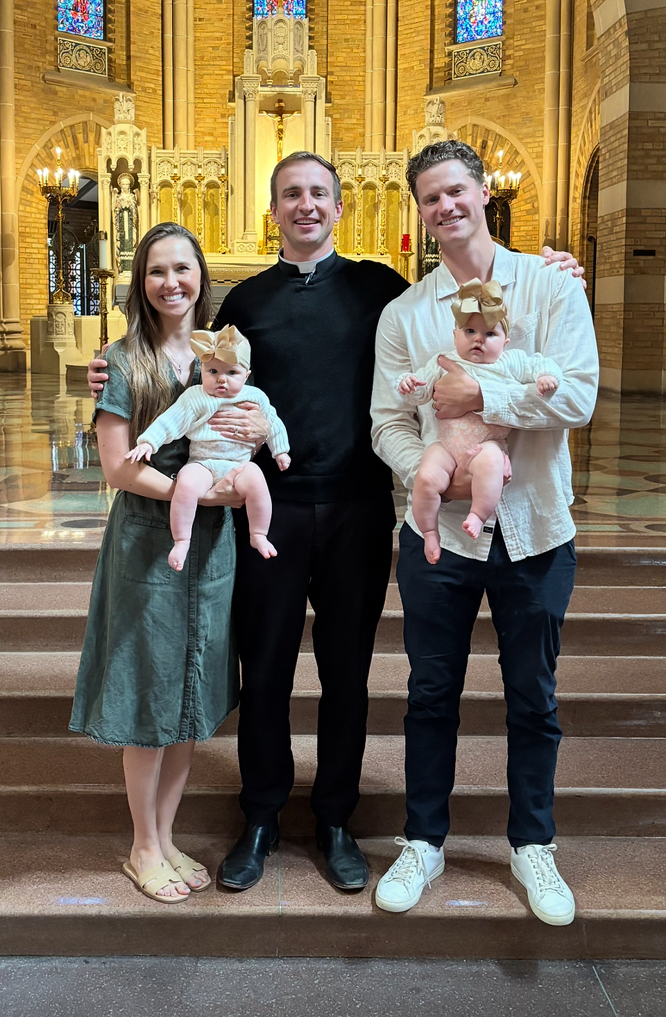 A couple holding babies with bows stands beside a priest inside a church with ornate golden decor and stained glass windows, all smiling.