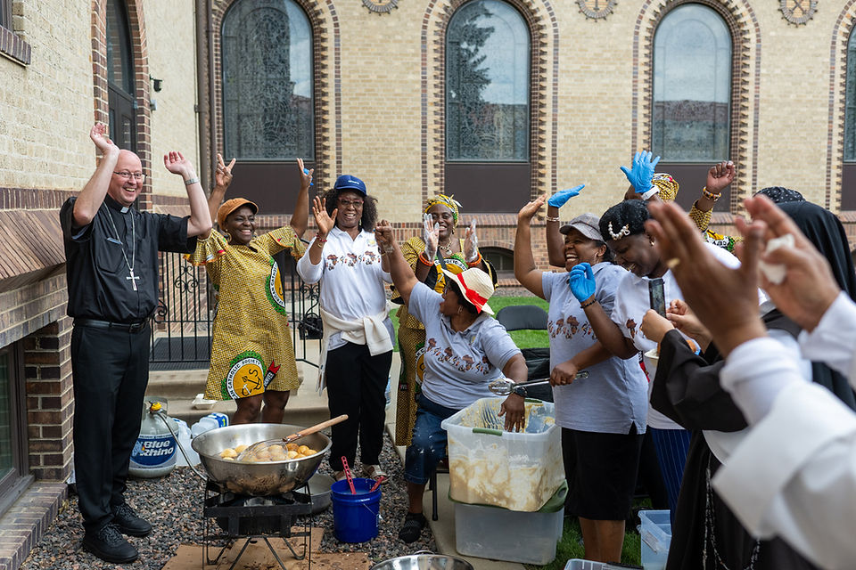 Joyful group, including a priest, raises hands and dances while cooking outdoors beside a church building.