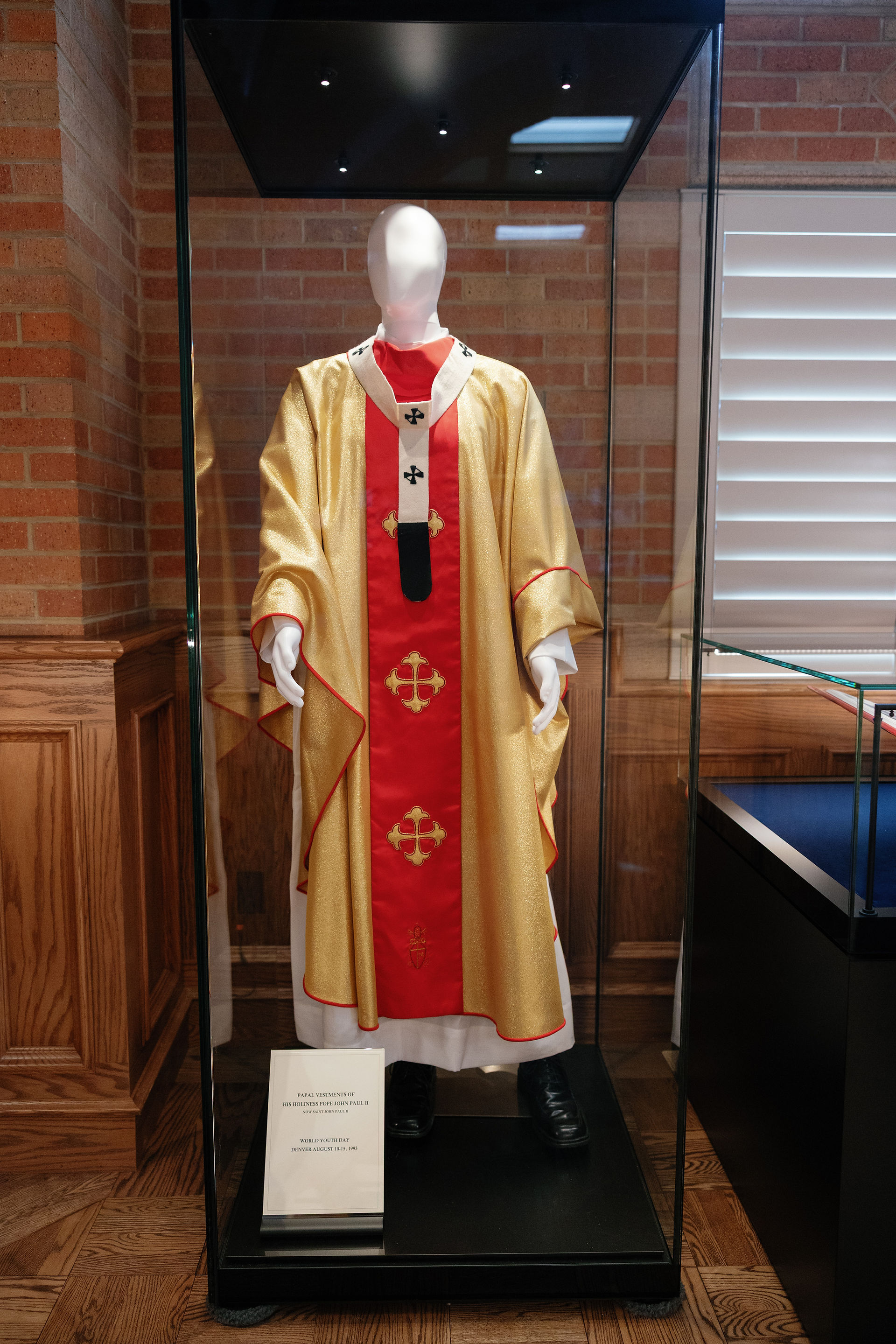 Mannequin in papal vestments of gold and red with crosses, displayed in a glass case against a brick wall, with a description plaque.