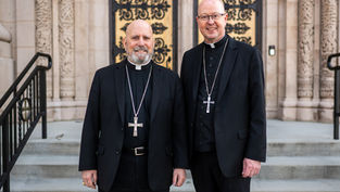 Two clergy members in black attire with cross necklaces stand outside an ornate building, smiling. Stone steps and decorative patterns in background.