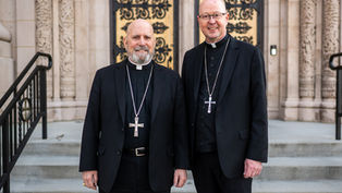 Two clergy members in black attire with cross necklaces stand outside an ornate building, smiling. Stone steps and decorative patterns in background.