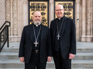 Two clergy members in black attire with cross necklaces stand outside an ornate building, smiling. Stone steps and decorative patterns in background.
