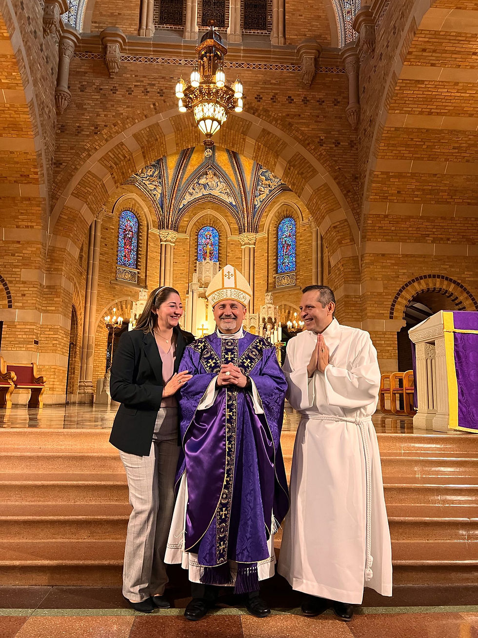 Three people stand in a cathedral-style church, smiling. The central figure in purple robes and a hat is flanked by a woman in a suit and a man in white.