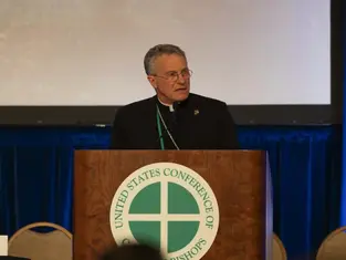 A man speaks at a podium labeled "United States Conference of Catholic Bishops." Blue backdrop, serious expression, wearing clerical attire.