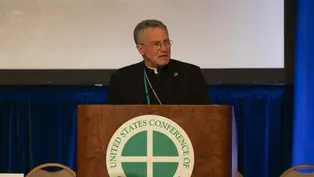A man speaks at a podium labeled "United States Conference of Catholic Bishops." Blue backdrop, serious expression, wearing clerical attire.