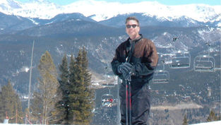 Smiling person in ski gear holding poles on a snowy mountain with evergreen trees, ski lifts, and snowy peaks in the background. Sunny day.