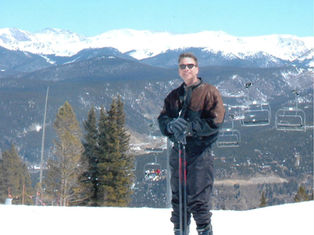 Smiling person in ski gear holding poles on a snowy mountain with evergreen trees, ski lifts, and snowy peaks in the background. Sunny day.