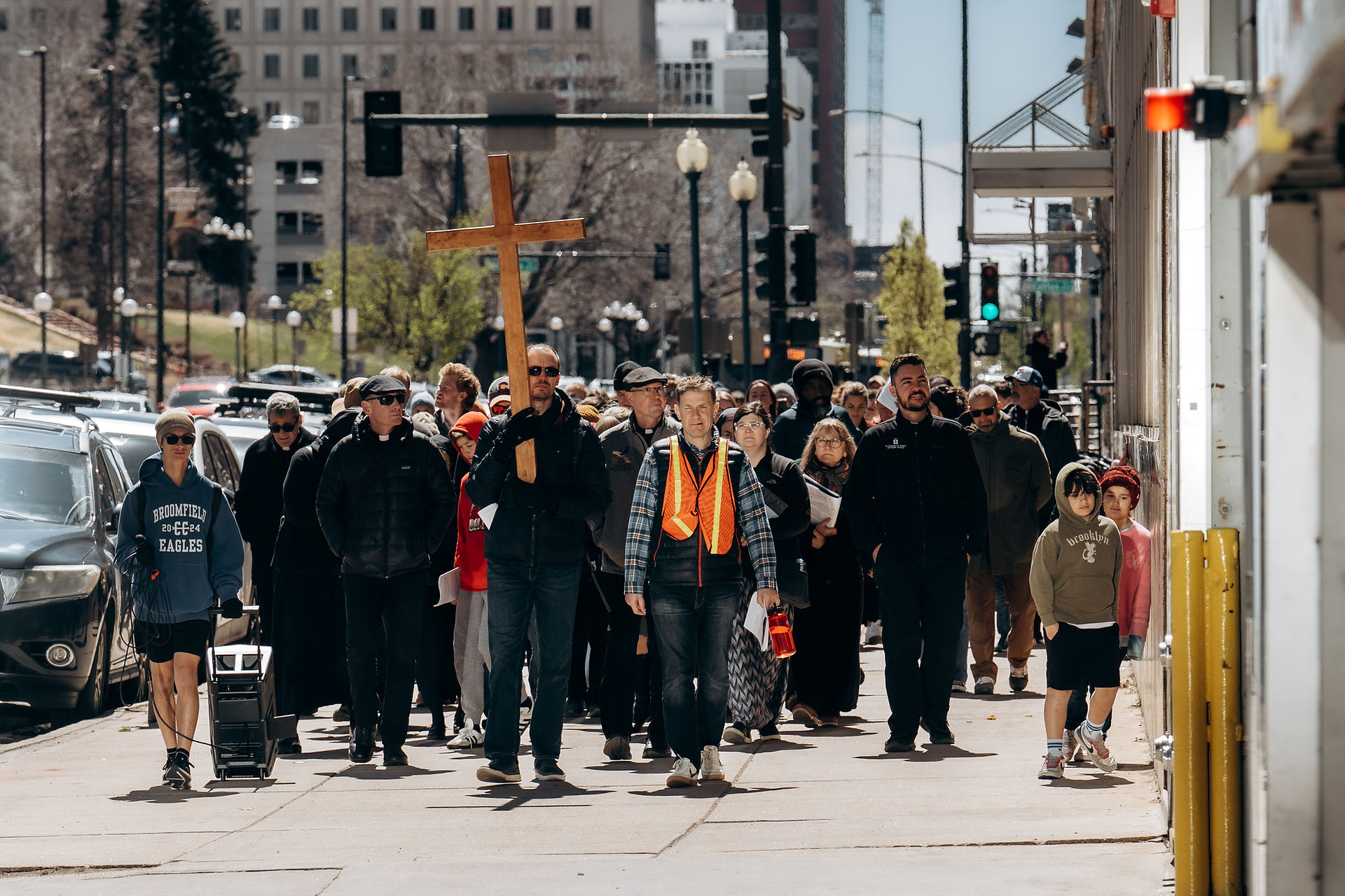 A large group walks together along a city street, following a wooden cross during a Way of the Cross procession.