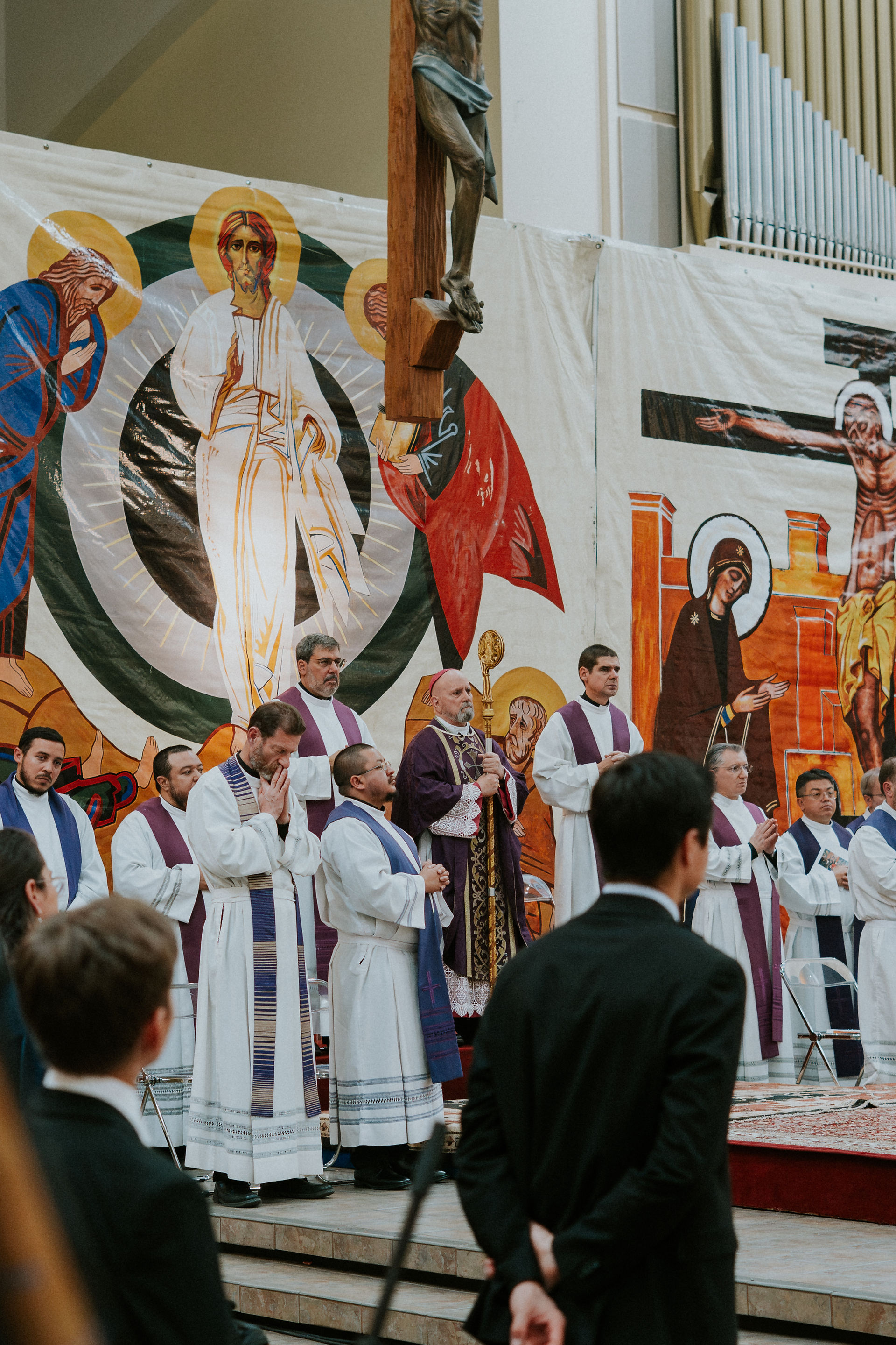 Clergy in white and purple robes participate in a religious ceremony before a colorful icon backdrop, with a large crucifix above.