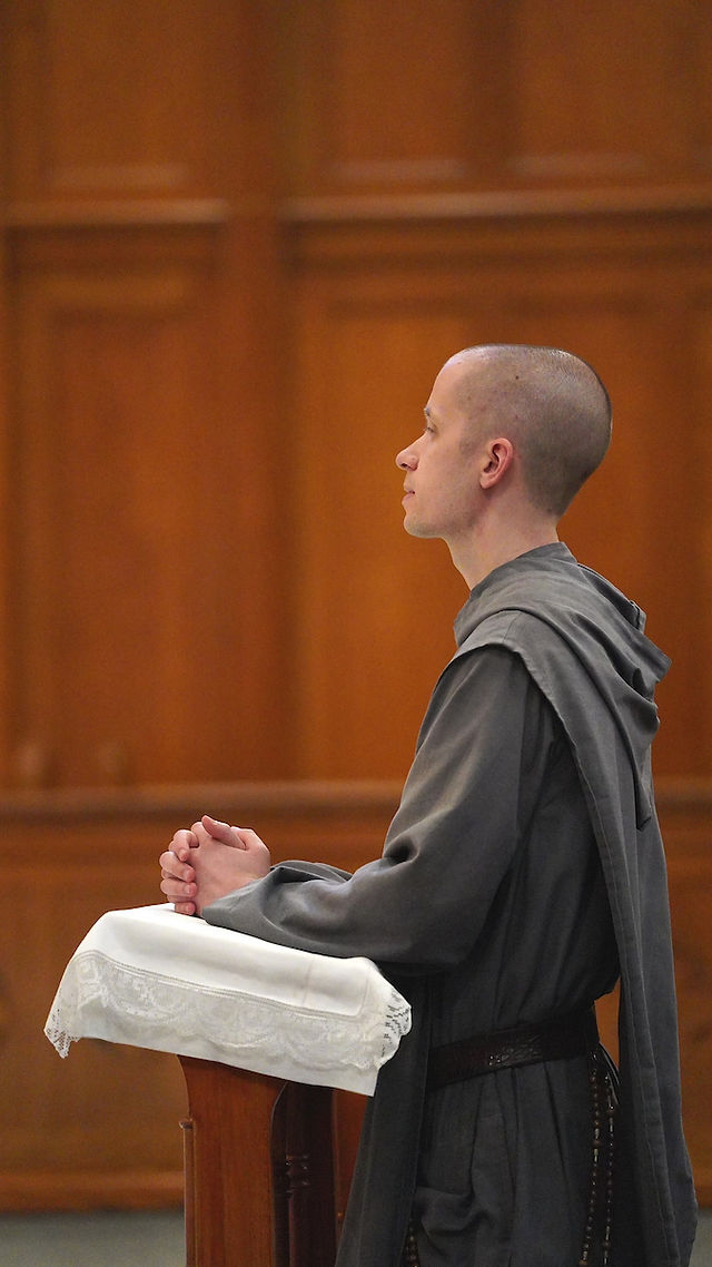 A person in a gray robe prays at a wooden altar, hands clasped, in a serene church setting with soft lighting.