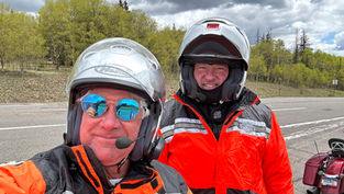 Two people in helmets and orange jackets pose by a road, under cloudy skies. One wears reflective sunglasses. Trees and a motorcycle are in the background.