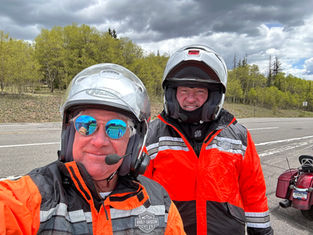 Two people in helmets and orange jackets pose by a road, under cloudy skies. One wears reflective sunglasses. Trees and a motorcycle are in the background.