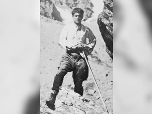 Man in period clothing leans on a walking stick in rocky terrain. He looks determined, set against a rugged mountain backdrop. Black and white photo.