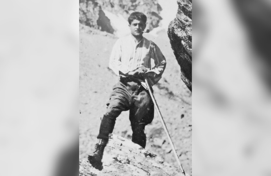 Man in period clothing leans on a walking stick in rocky terrain. He looks determined, set against a rugged mountain backdrop. Black and white photo.