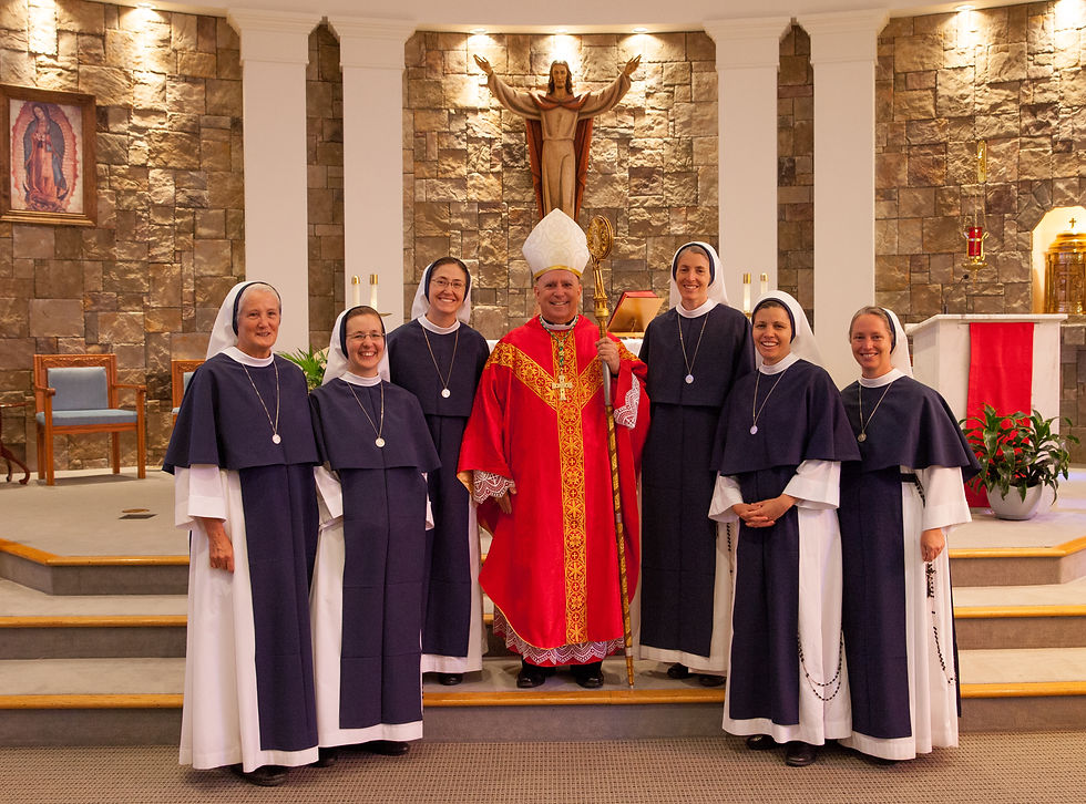 A bishop in red robes stands with smiling nuns in dark blue habits in a church, with a stone wall and religious statue behind them.