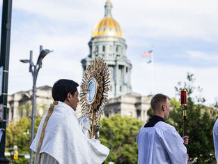 Catholic procession with priests carrying a gold monstrance and red candle before a domed capitol building under a bright sky.