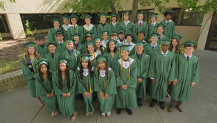 Graduates in green caps and gowns pose outdoors by a building. They're smiling, exuding a joyful, celebratory mood under a leafy tree.
