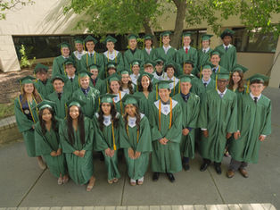 Graduates in green caps and gowns pose outdoors by a building. They're smiling, exuding a joyful, celebratory mood under a leafy tree.