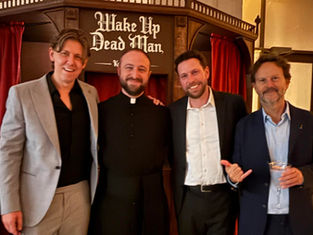 Four men in suits stand smiling in a room with red curtains. One wears a clerical collar. Sign reads "Wake Up Dead Man."