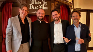 Four men in suits stand smiling in a room with red curtains. One wears a clerical collar. Sign reads "Wake Up Dead Man."