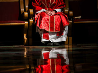 Person in red ornate robe sits in a wooden chair, hands clasped. Floor reflection visible. Calm mood, no text.