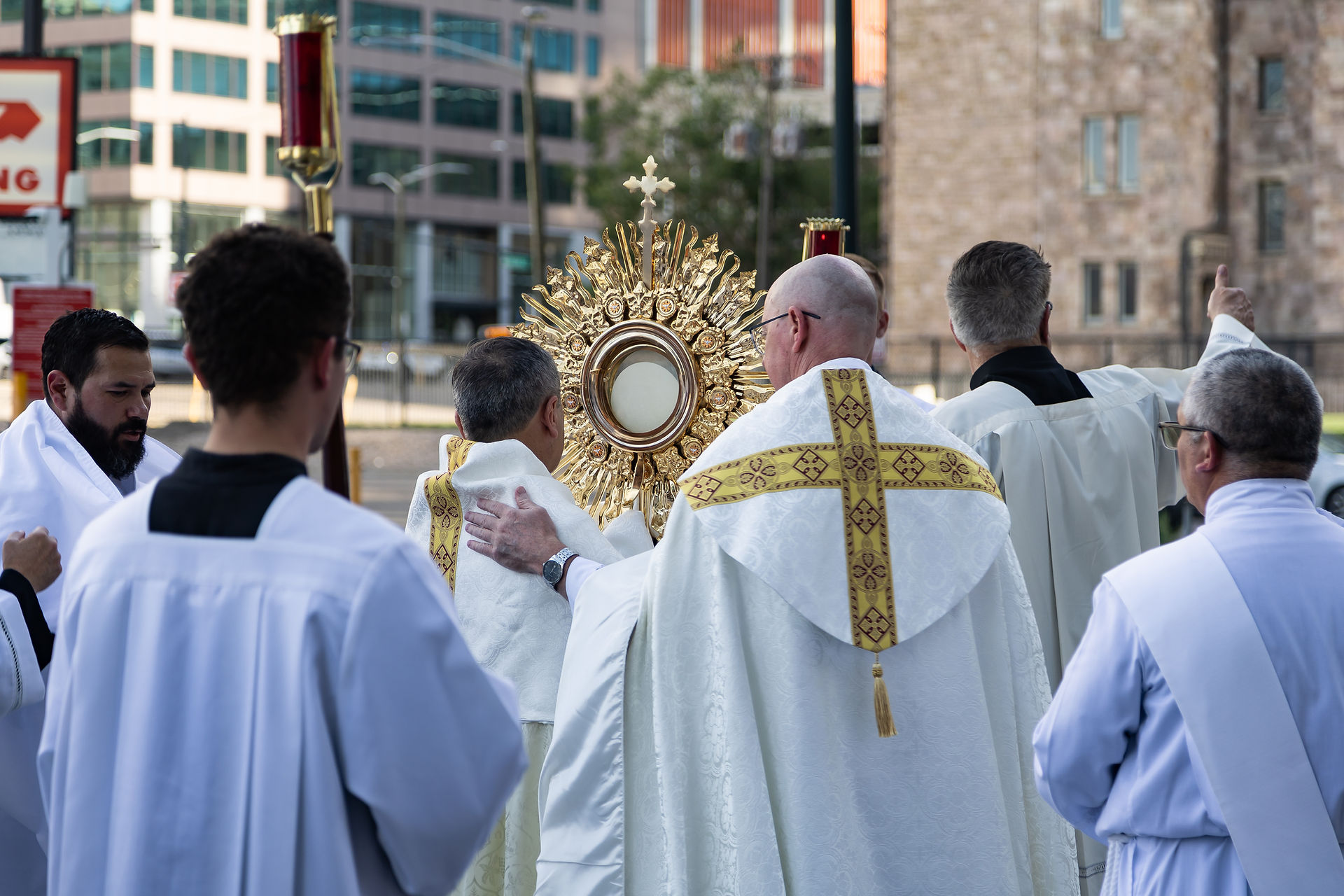 Catholic priests in white vestments process behind a gold monstrance on a city street, a solemn outdoor ritual