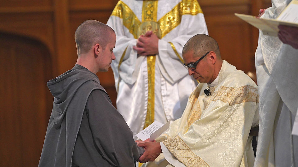 A monk kneels, holding hands with a seated priest in ornate robes, during a solemn ceremony inside a church.