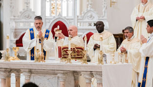 Priests in white robes participate in a religious service at an ornate altar, holding a chalice. The setting is solemn and reverent.