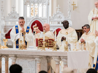 Priests in white robes participate in a religious service at an ornate altar, holding a chalice. The setting is solemn and reverent.