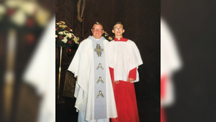 Priest in white robe with cross stands next to altar boy in red and white gown. They are in a church, with flowers and a crucifix behind them.