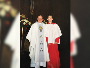 Priest in white robe with cross stands next to altar boy in red and white gown. They are in a church, with flowers and a crucifix behind them.