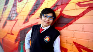 Boy with blue glasses leans against colorful graffiti wall. Wears a school uniform featuring a red tie and a logo. Smiling confidently.