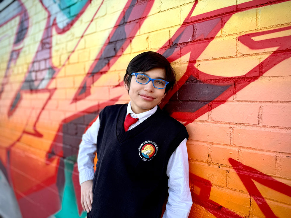 Boy with blue glasses leans against colorful graffiti wall. Wears a school uniform featuring a red tie and a logo. Smiling confidently.