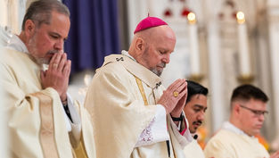 Clergymen in white robes, heads bowed in prayer, stand in a church with intricate arches. A man wears a pink skullcap, setting is solemn.