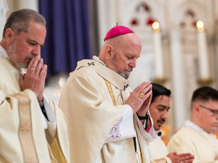 Clergymen in white robes, heads bowed in prayer, stand in a church with intricate arches. A man wears a pink skullcap, setting is solemn.