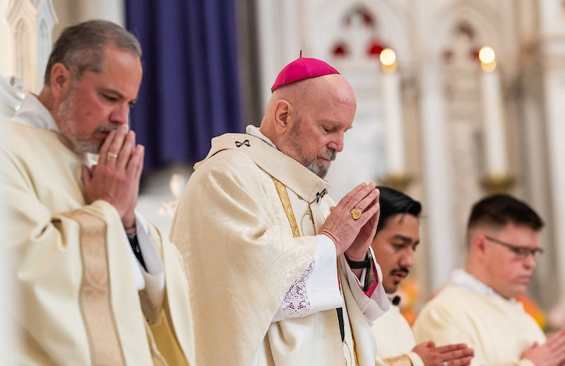 Clergymen in white robes, heads bowed in prayer, stand in a church with intricate arches. A man wears a pink skullcap, setting is solemn.