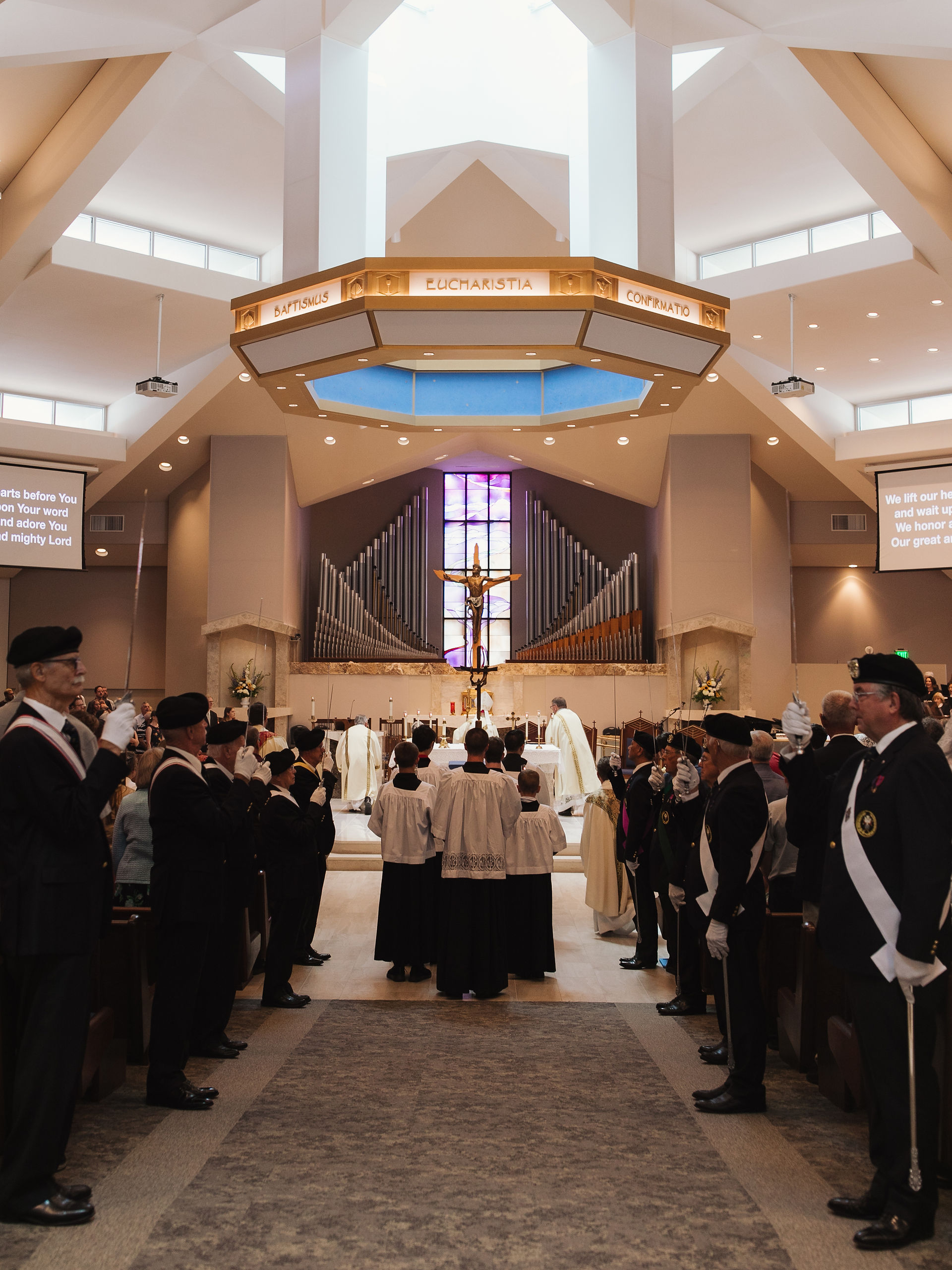 Interior of a church with people in ceremonial attire. Central altar with crucifix, organ pipes, and stained glass. Words "Eucharistia" visible.
