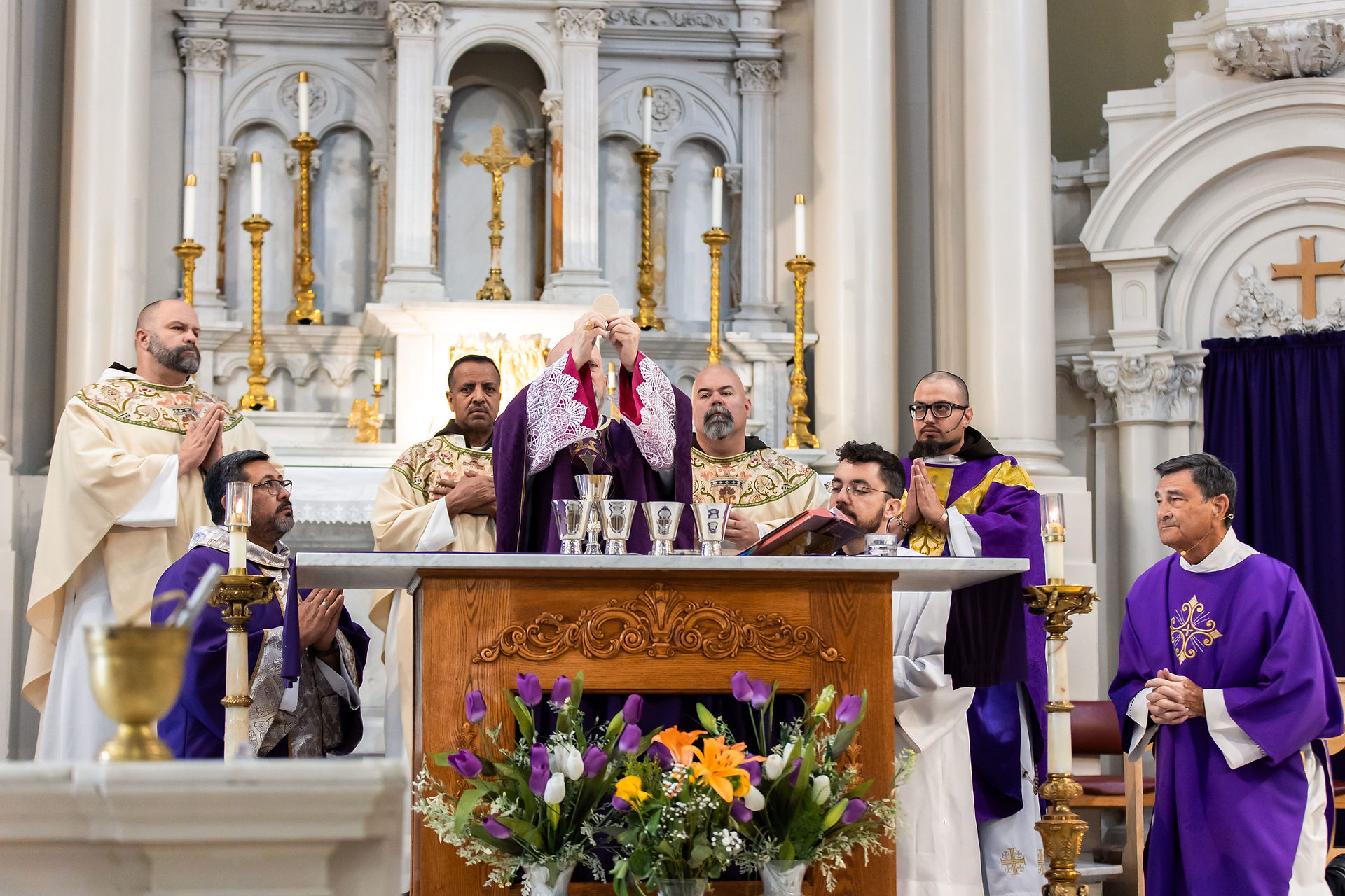 Clergy in ornate robes perform a Eucharist ceremony at an ornate altar, with flowers and candles, in a bright church.