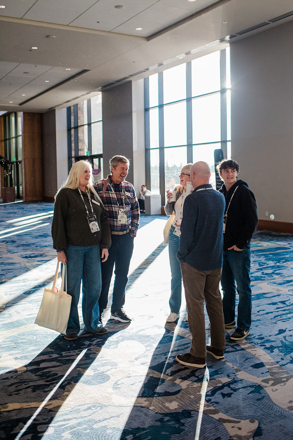 Group of conference attendees wearing name badges stand in a sunlit hallway, talking and smiling together near large windows.