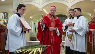 A religious ceremony with clergy in red and white robes; a man sprinkles holy water over palm fronds. Elaborate arches in the background.
